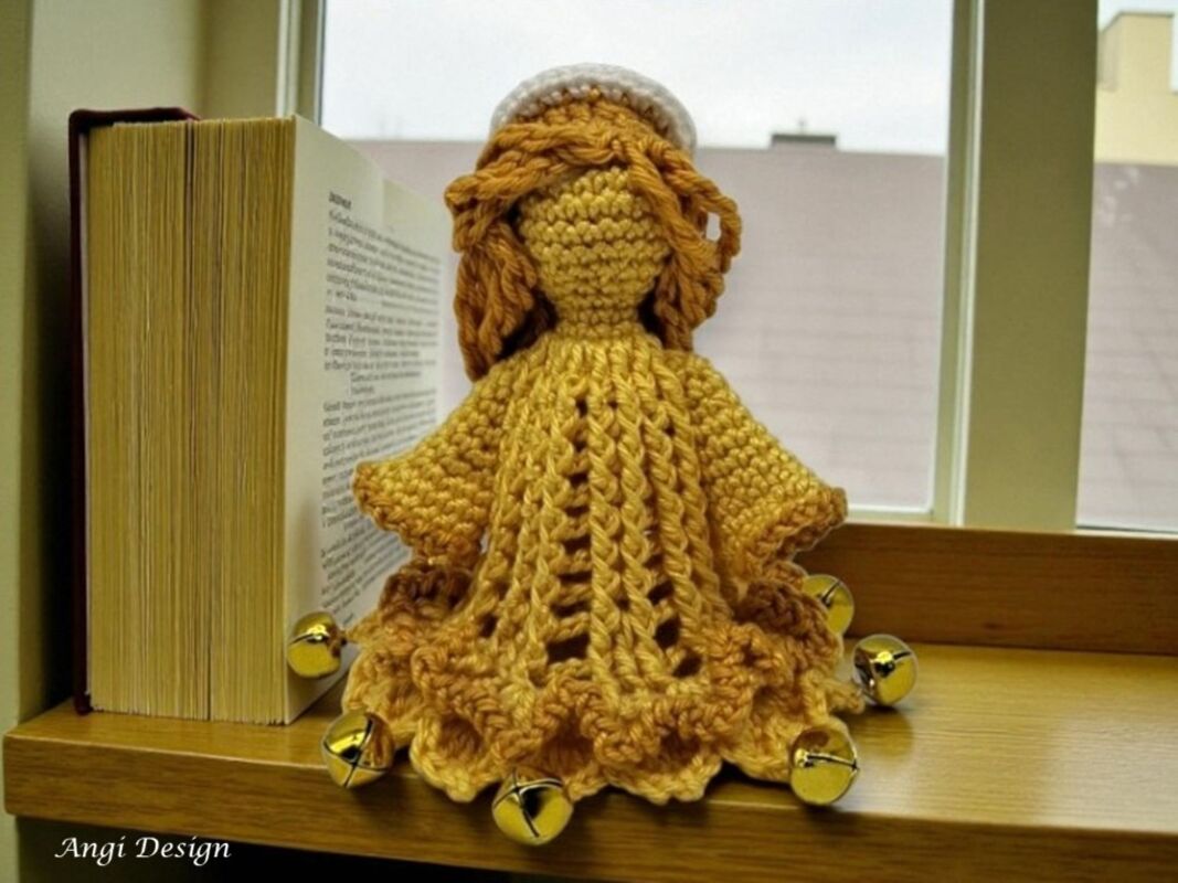 Gehäkeltes Engel-Amigurumi in senfgelbem Garn mit Rüschenrock und goldenen Glöckchen auf Fensterbank neben Buch