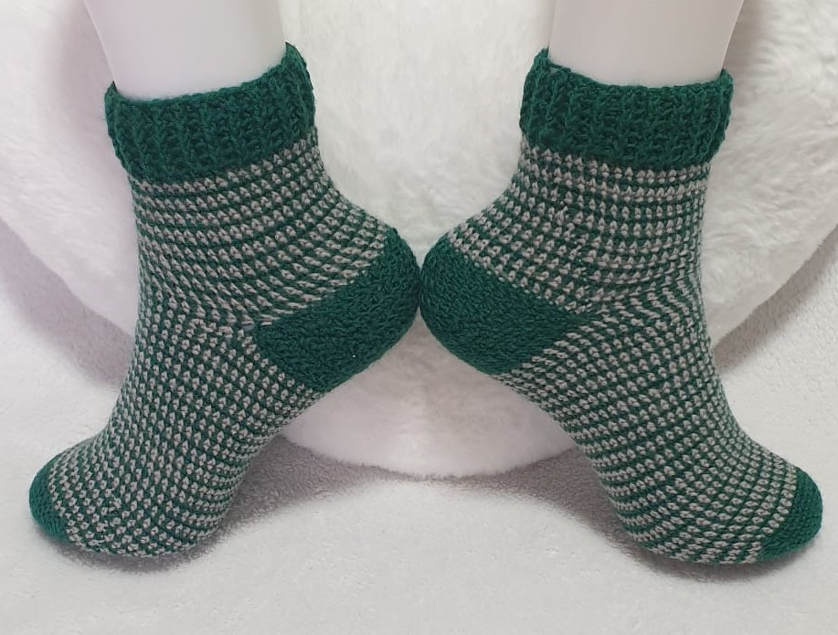 Paar gehäkelter Knöchelsocken in Grün und Hellgrau mit gemustertem Schaft und einfarbigen Bündchen, Fersen und Spitzen auf weißem Untergrund.