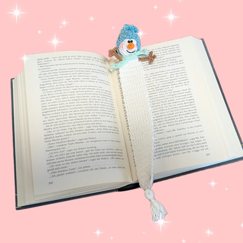 Gehäkeltes Schneemann-Lesezeichen mit blauer Mütze und mintgrünem Schal in einem geöffneten Buch.