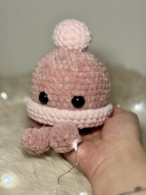 Rosa gehäkelter Amigurumi-Oktopus mit schwarzen Sicherheitsaugen und Pompons, in einer Hand auf flauschigem Untergrund vor Lichterkette gehalten.