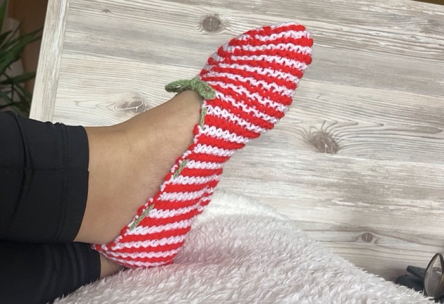 Fuß mit gestricktem rot-weiß gestreiftem Slipper und grüner Schleife auf weißem, flauschigem Untergrund.