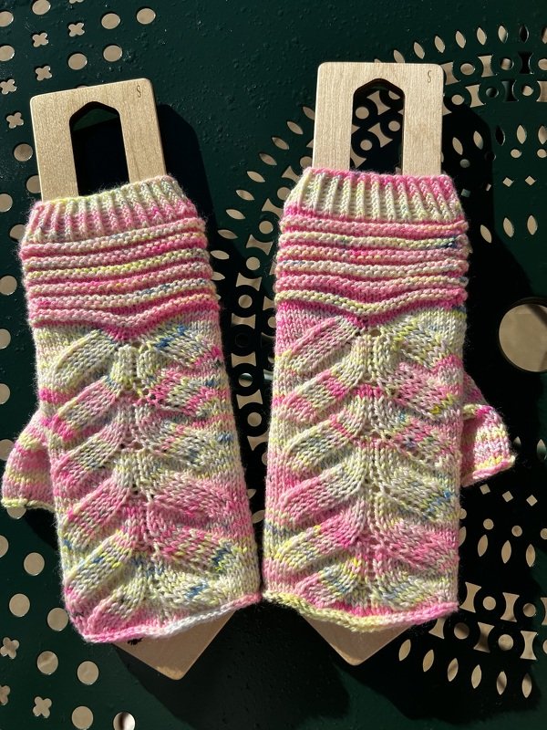 Ein Paar gestrickter, fingerloser Armstulpen in Rosa-Gelb-Melange mit Blattmuster auf Holzblockern.
