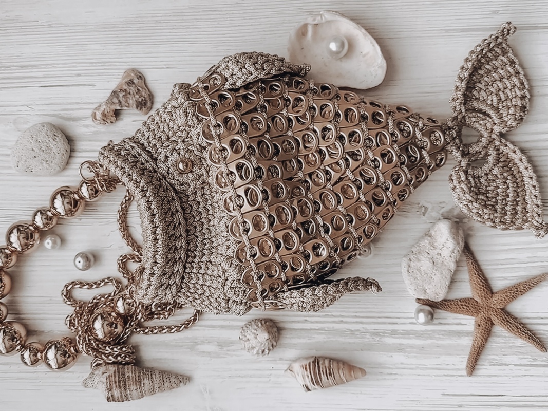 Beige hand-crocheted fish bag with ring-pull tab scales and beaded-chain strap on a light wooden surface with shells