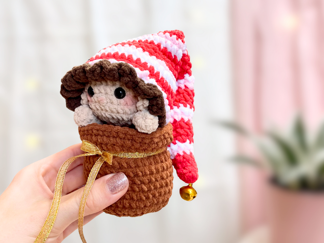Hand hält gehäkelten Amigurumi-Wichtel mit rot-weiß gestreifter Mütze und Glöckchen in braunem Beutel mit Goldband.