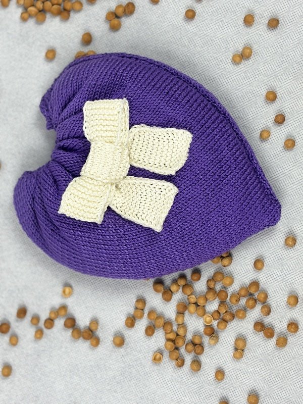 Purple knitted heart pillow with a cream bow on a light gray surface with scattered brown beads.