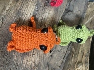 Zwei handgehäkelte Amigurumi‑Tierchen in Orange und Grün mit schwarzen Augen auf Holz.