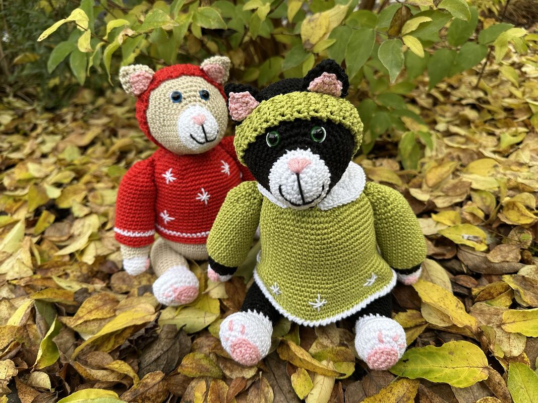 Zwei gehäkelte Katzen-Amigurumis sitzen auf Herbstblättern; eine beige mit rotem Pullover, die andere schwarz-weiß mit grünem Pullover und Mütze.