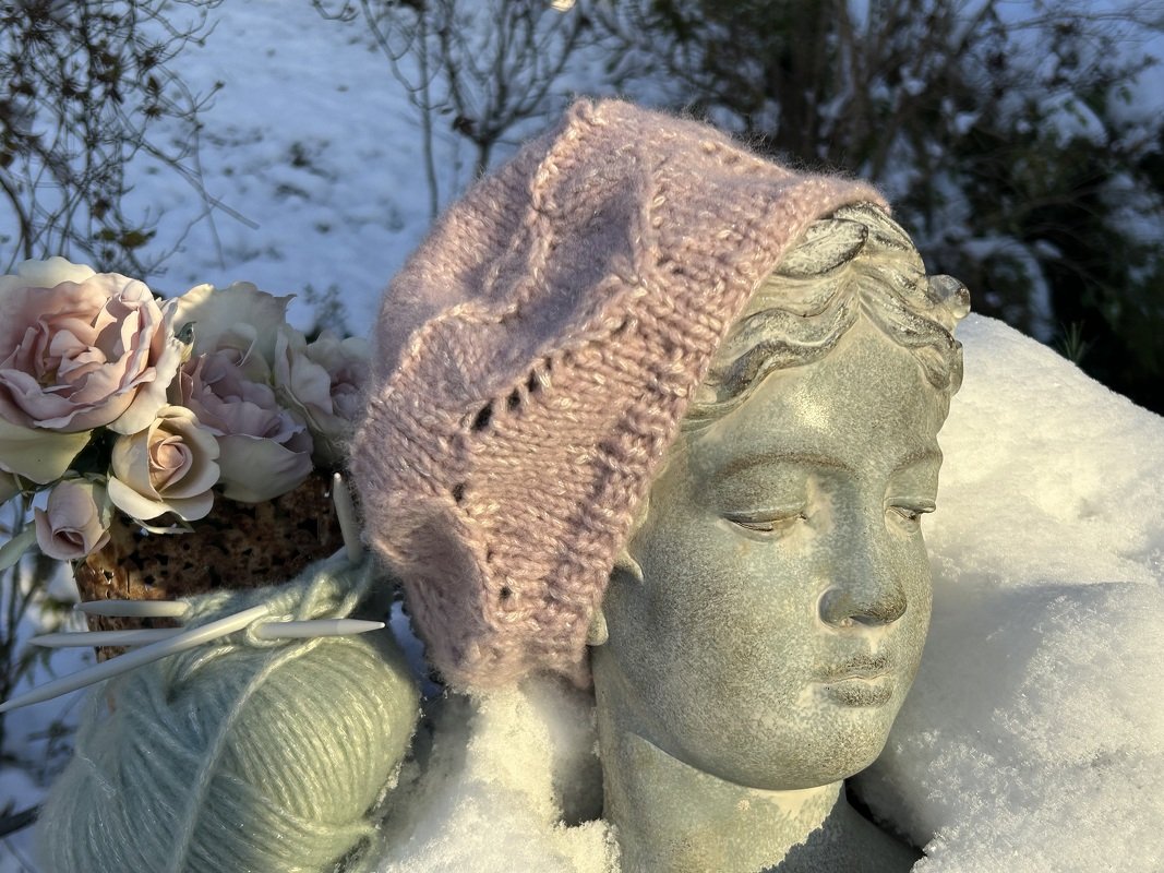 Zartrosé gestrickte Baskenmütze auf einer steinernen Büste im Schnee, mit Garn und Stricknadeln.