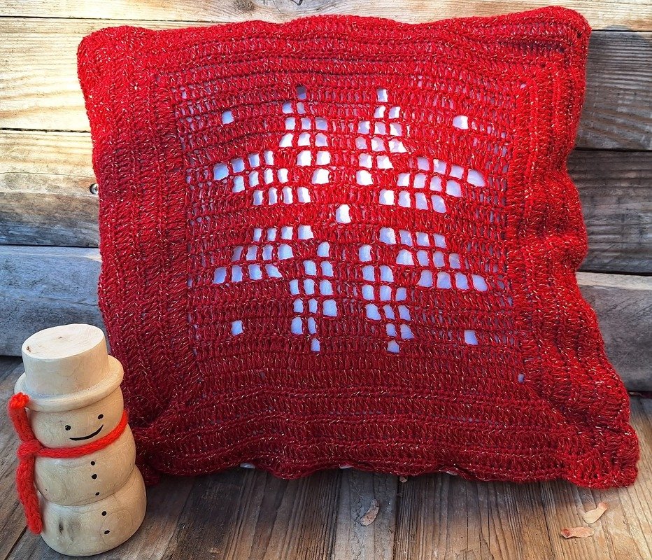 Rotes gehäkeltes Kissen mit durchbrochenem Schneeflockenmuster und kleiner Holzfigur auf Holzuntergrund.