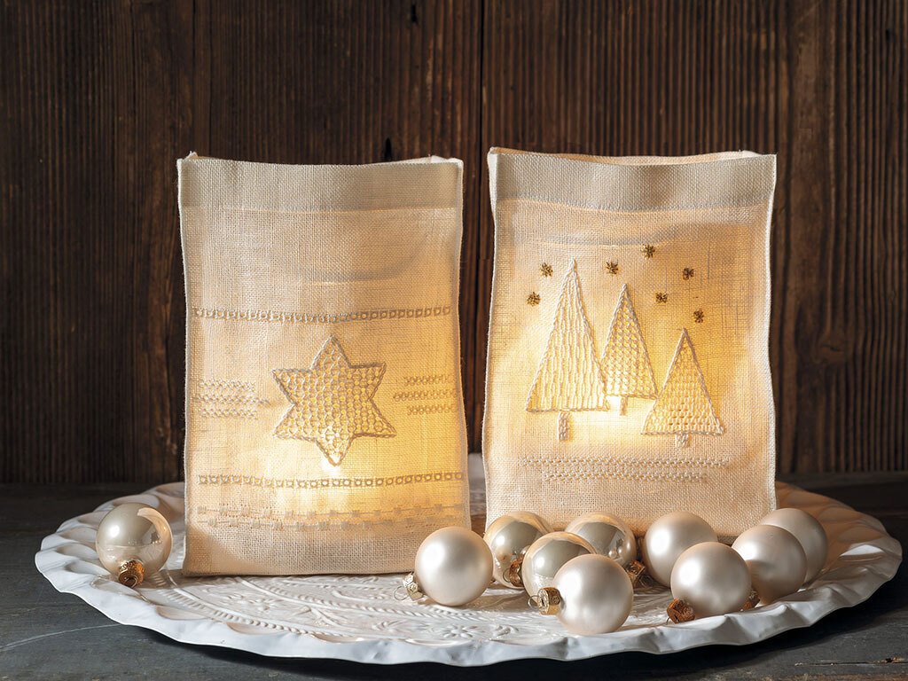 Zwei leuchtende Stofflaternen mit besticktem Stern und Tannenbäumen, umgeben von perlweißen Weihnachtskugeln auf einem Dekoteller vor einer Holzwand.