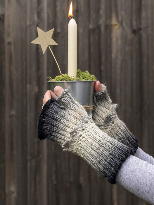 Hände mit gestrickten fingerlosen Armstulpen im Ombre‑Grau halten einen kleinen Topf mit brennender Kerze