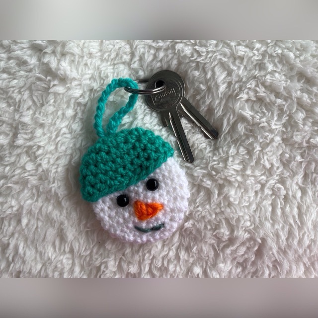Gehäkelter Schneemann-Schlüsselanhänger mit türkisfarbener Mütze auf weißem, flauschigem Untergrund an einem Schlüsselring.