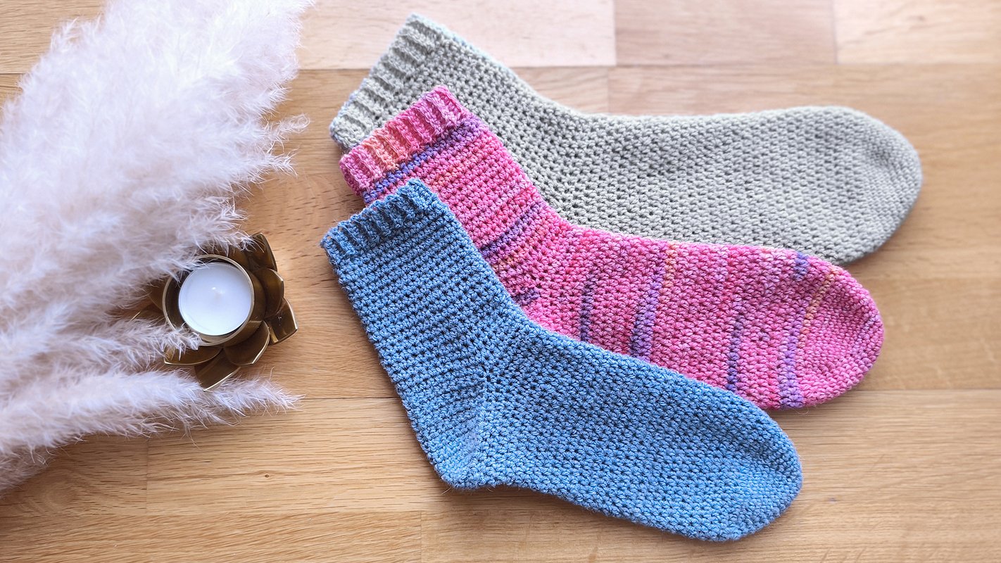 Drei gehäkelte Socken in Beige, Rosa und Blau auf Holzbrett neben Kerze und Pampasgras