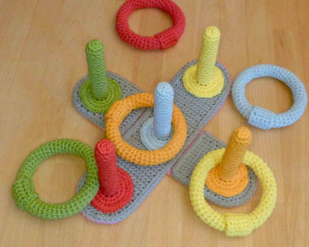 Nahaufnahme eines gehäkelten Ringwurfspiels auf einem Holzboden mit farbigen Stäben und Ringen in Grün, Gelb, Orange, Hellblau und Rot.