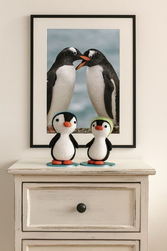 Zwei gehäkelte Amigurumi-Pinguine mit blauer und grüner Mütze auf einer Kommode vor gerahmtem Pinguinfoto.