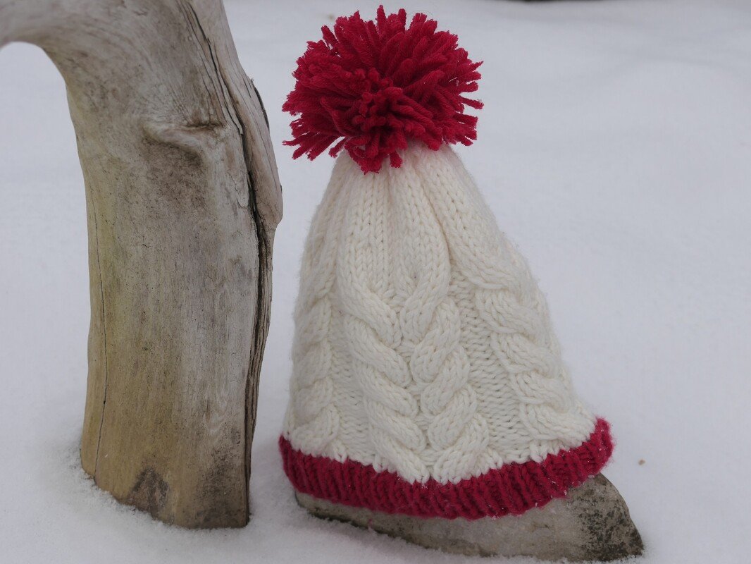 nahaufnahme weiße gestrickte Zopfmütze mit rotem geripptem Rand und rotem Bommel auf Schnee
