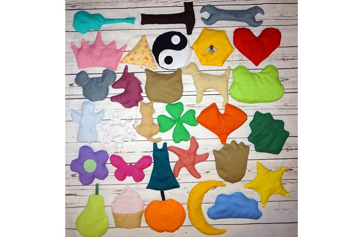Verschiedene genähte und gestopfte Stoffformen in unterschiedlichen Farben und Motiven wie Tiere, Früchte und Werkzeuge auf einem Holztisch ausgelegt.