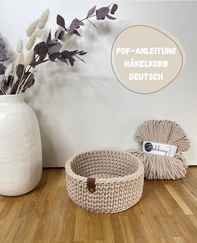 Beiger, runder Häkelkorb aus dickem Garn auf Holzfußboden neben Knäuel und Vase
