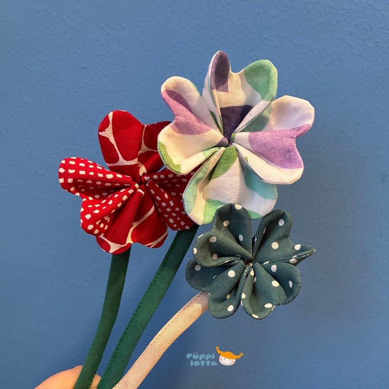 Drei genähte Stoffblüten in Rot, Pastell und Grün an bezogenen Stielen vor blauem Hintergrund