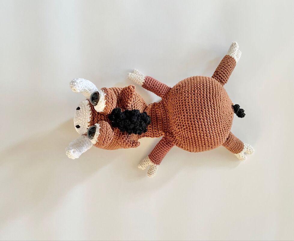 Gehäkeltes braunes Amigurumi-Rind mit weißen Hörnern und schwarzem Nackenbesatz, liegend