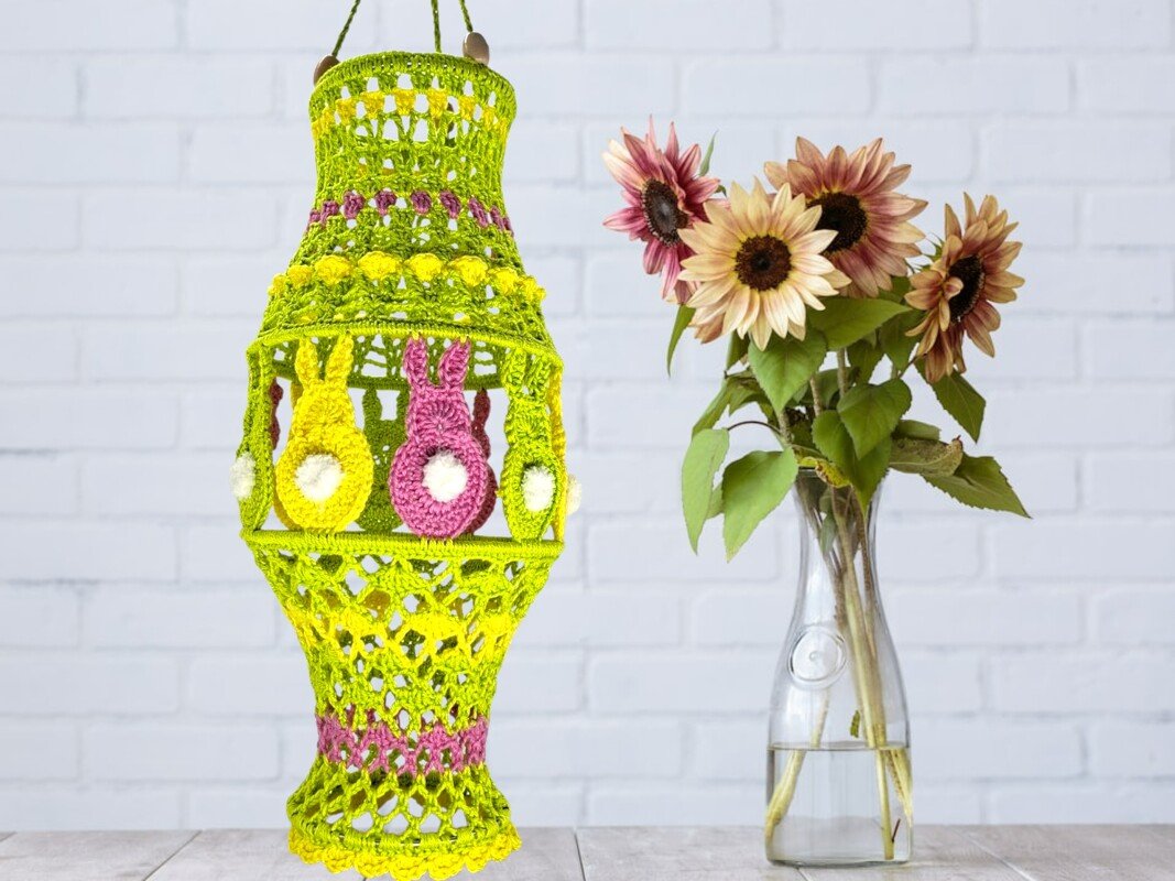 Gehäkelte grüne Laterne mit rosa und gelben Hasen-Applikationen und weißen Bommelschwänzchen neben einer Vase mit Sonnenblumen