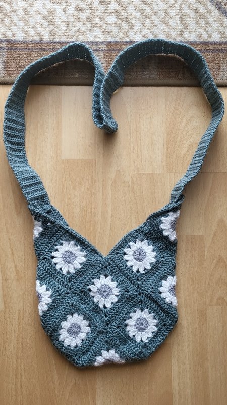 Gehäkelte Umhängetasche aus Granny
‑Squares mit weißen Blüten auf graugrünem Grund, langer Riemen liegt herzförmig auf Holzboden