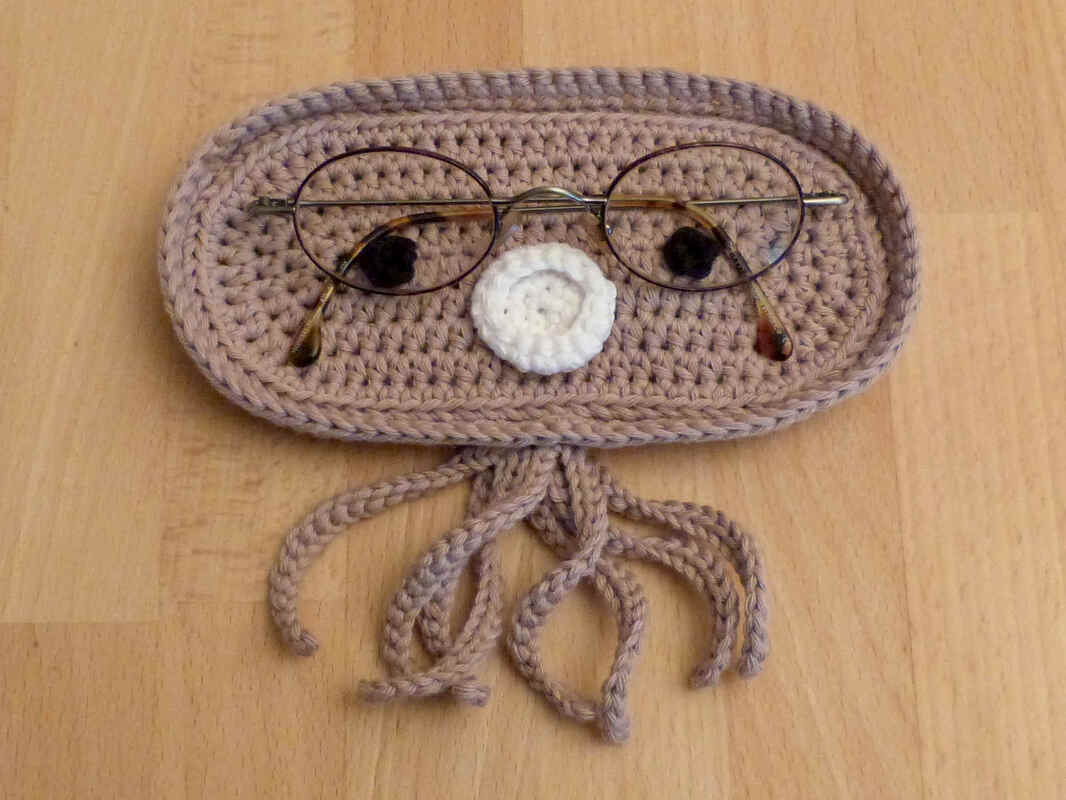 Kompaktes, ovales Häkelutensilo in Beige mit weißer Applikation und Quasten, Brille liegt darauf.