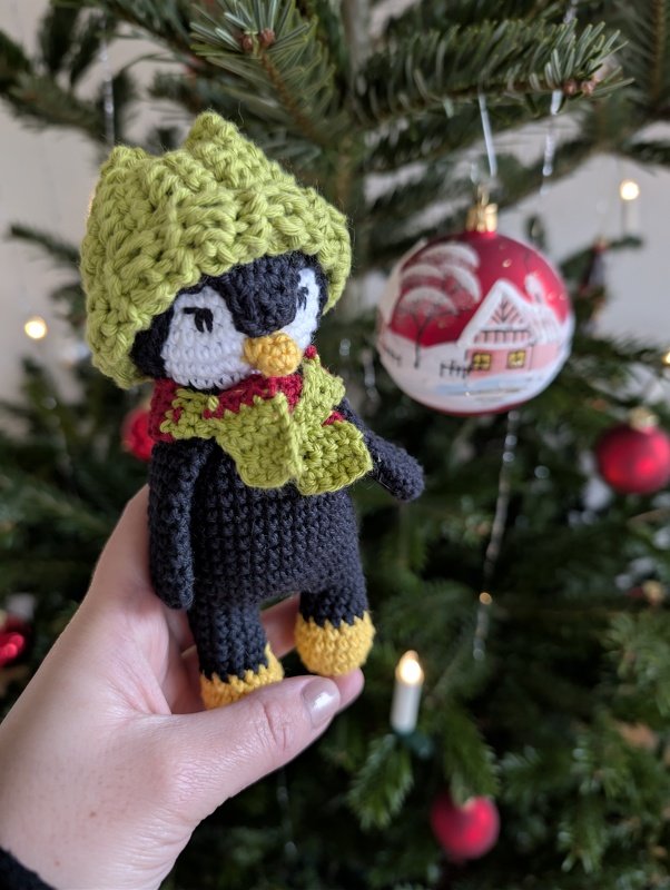 Gehäkelter Amigurumi-Pinguin mit grüner Mütze und Schal vor einem Weihnachtsbaum