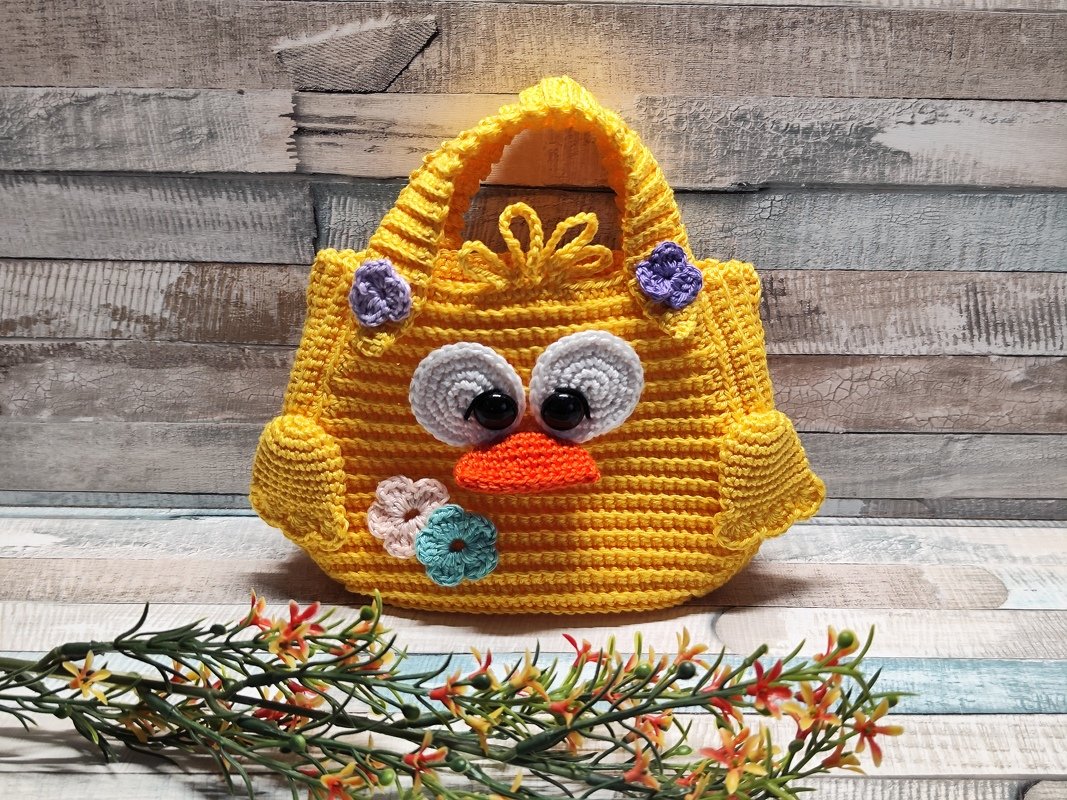 Gelbe gehäkelte Küken-Tasche mit großen Augen, orangefarbenem Schnabel und Blumenapplikationen vor Holzhintergrund.