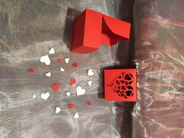 Rote Geschenkbox, rote Karte mit filigranem Herzausschnitt und verstreute rote und weiße Papierherzen auf glänzendem Untergrund.