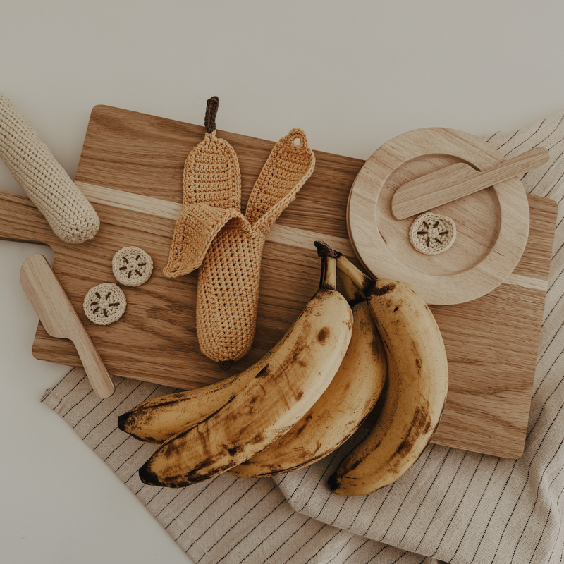 Teilweise geschälte, gehäkelte Banane mit drei gehäkelten Scheiben auf Holzbrett neben Holzbesteck und echten Bananen.
