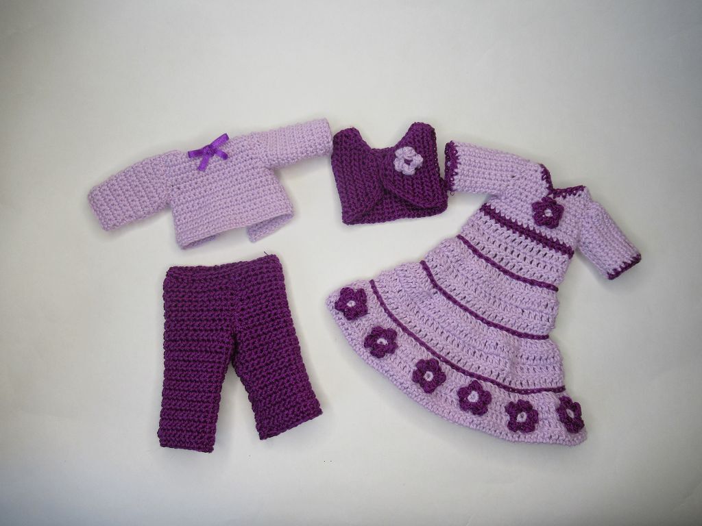 Vier gehäkelte Puppenkleidungsstücke in hellen und dunklen Lilatönen: Kleid mit Blüten, Hose, Bolero und Oberteil