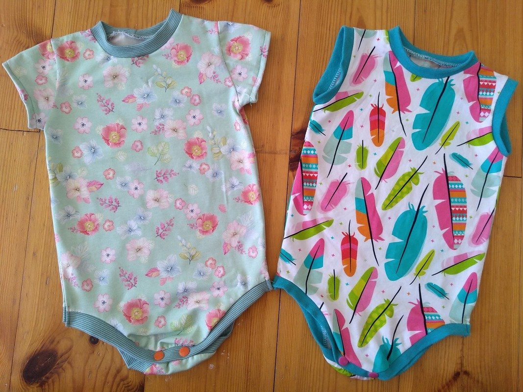 Zwei Babybodys auf Holz: mintgrüner Blumenbody kurzarm und weißer ärmelloser Body mit bunten Federn
