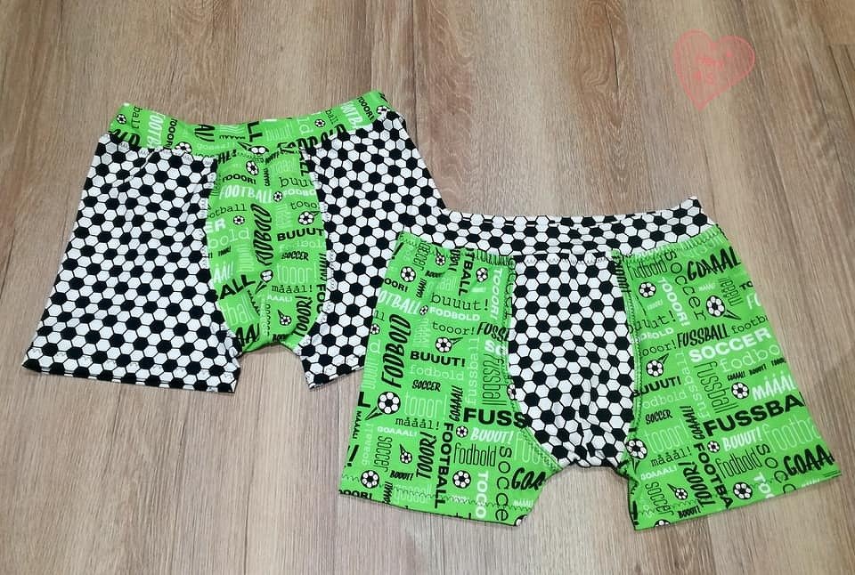 Zwei Kinder-Boxershorts mit schwarz-weißem Fußballmuster und grünen Einsätzen auf Holzboden