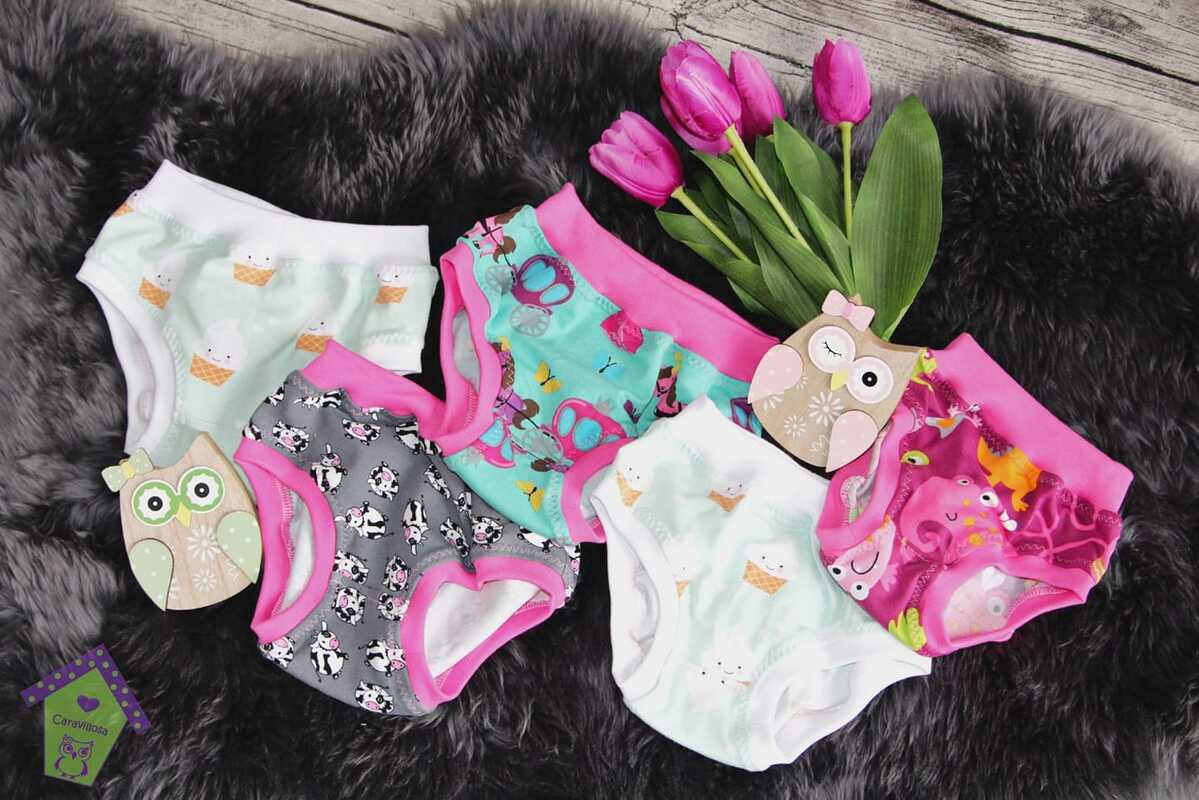 Bunte Baby- und Kinderunterhosen mit Eis-, Kuh-, Schmetterlings- und Meeresmotiven auf dunklem Kunstfell neben Holz-Eulen und pinken Tulpen.