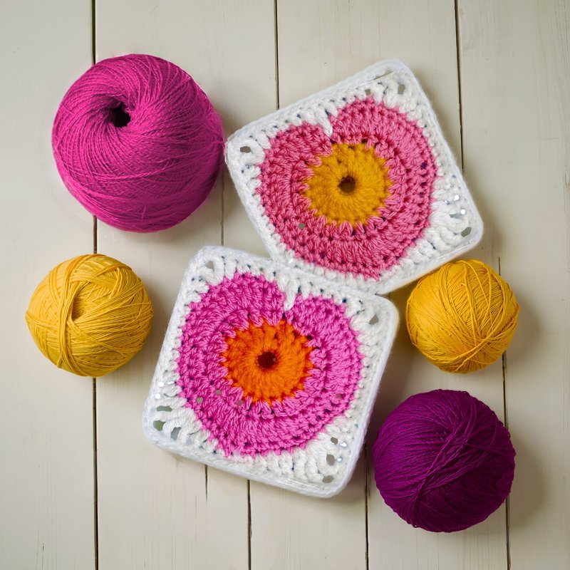 Two round-center granny square motifs in pink, orange, and white with four yarn balls on a light wood surface