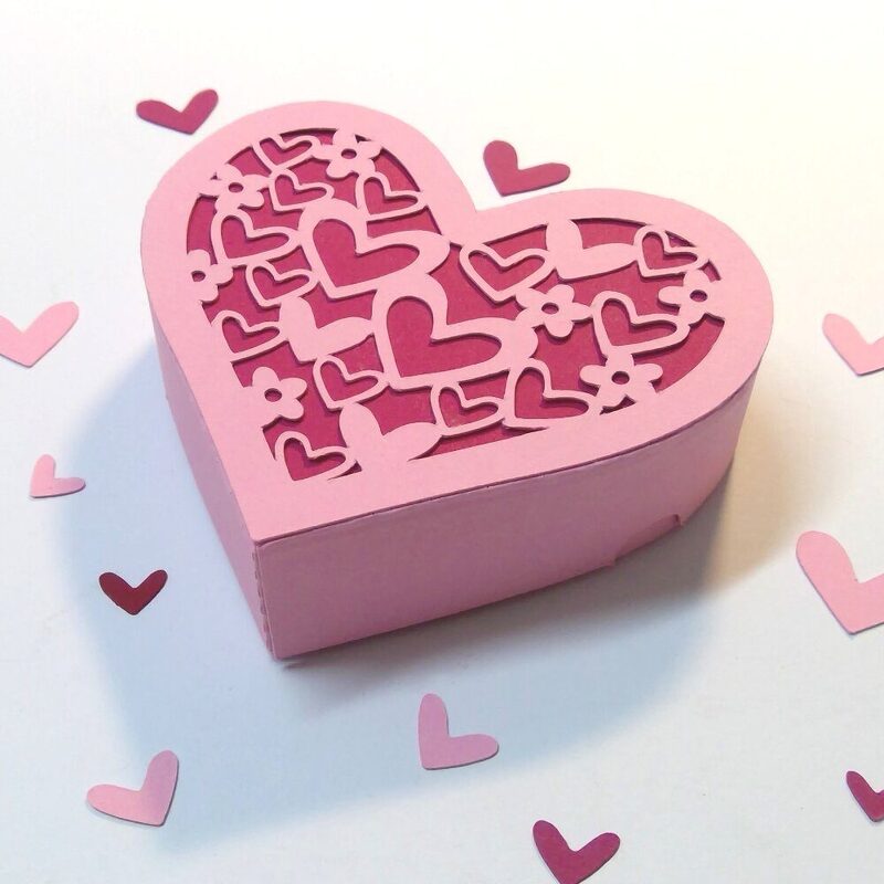 Rosa herzförmige Geschenkbox aus Papier mit fein ausgeschnittenem Deckelmotiv aus Herzen und Blumen, auf hellem Hintergrund mit verstreuten Papierherzen.