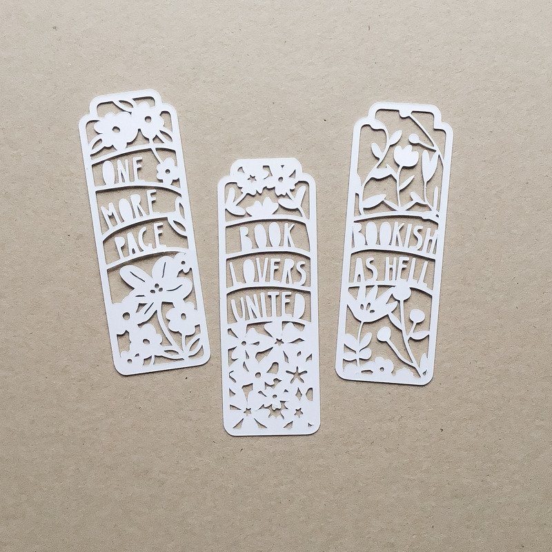Drei weiße, detailreich geplottete Lesezeichen aus Papier mit floralen Elementen und englischen Phrasen auf beigem Hintergrund.