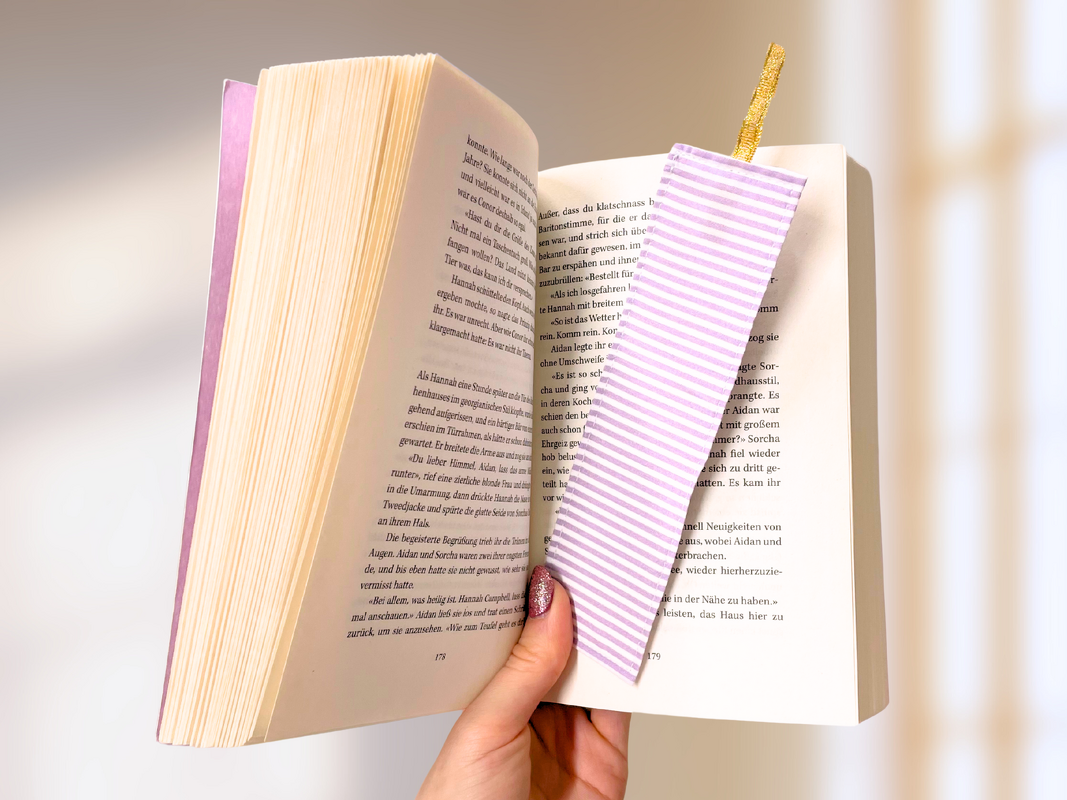 Genähtes Stofflesezeichen gestreift in helllila mit goldfarbener Schlaufe zwischen den Seiten eines Buchs