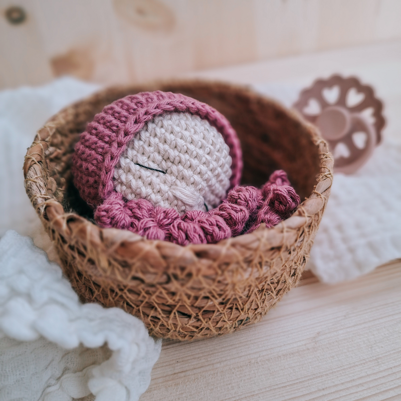 Gehäkeltes Baby mit rosa Mütze und Decke, liegend in einem geflochtenen Korb auf hellem Holzhintergrund.