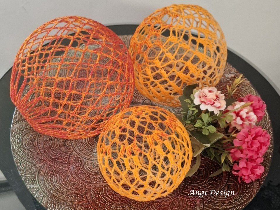 Drei gehäkelte offene Kugellampions in Gelb- und Orange-Tönen auf verzierter Platte neben Blumendekor