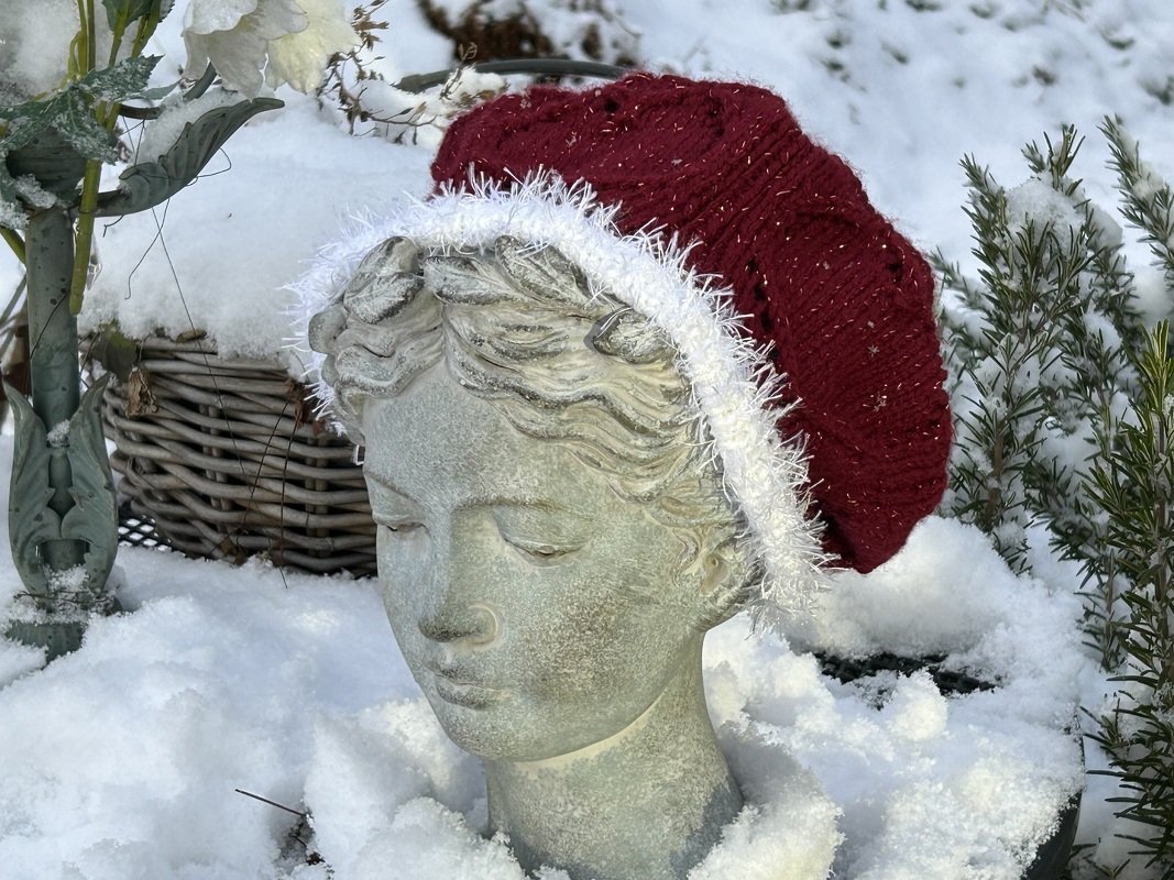 Dunkelrote Strickmütze mit weißem Flauschrand auf steinerner Büste im Schnee