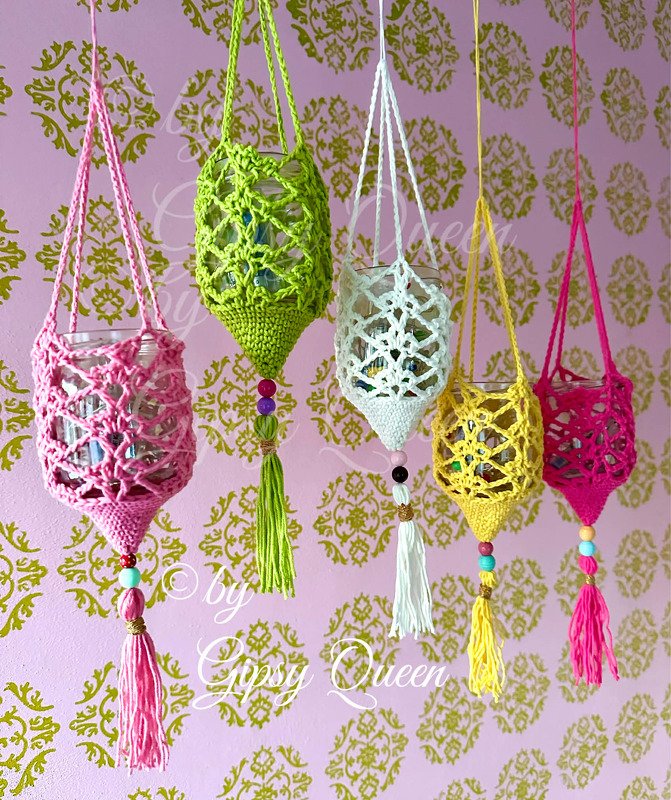 Fünf bunte gehäkelte Hüllen für Teelichtgläser mit Perlen und Quasten, hängend vor rosa Wand mit goldfarbenem Muster.