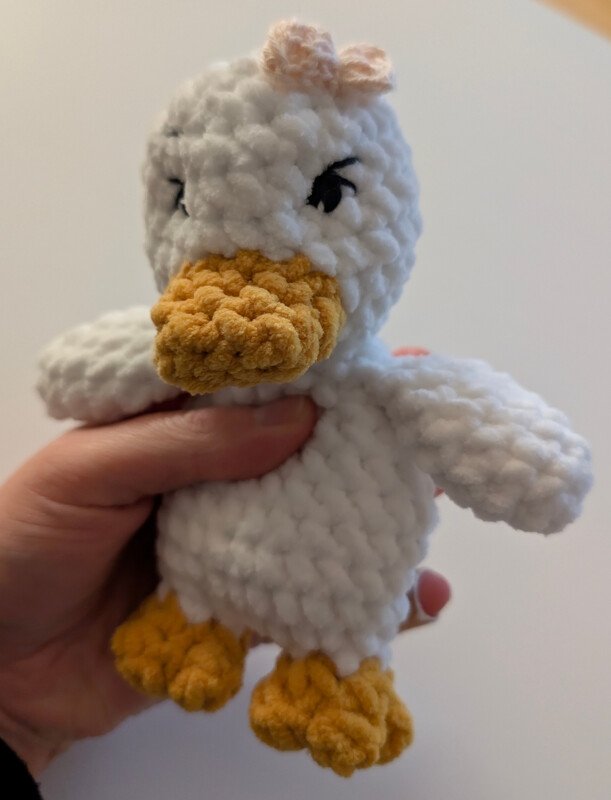Weißes gehäkeltes Enten-Amigurumi mit orangefarbenem Schnabel und Füßen und einer rosa Schleife, in einer Hand gehalten.
