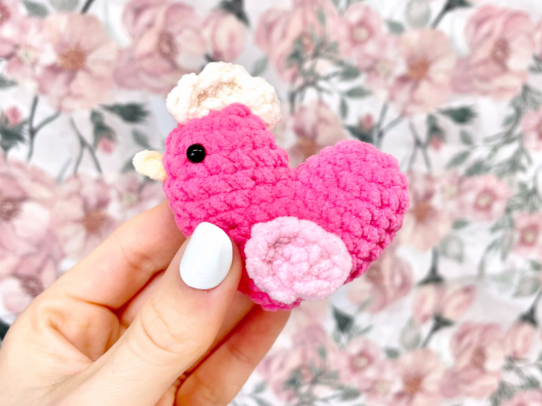 Hand mit weißen Nägeln hält kleines pinkes gehäkeltes Herz-Vogel-Amigurumi vor floralem Hintergrund