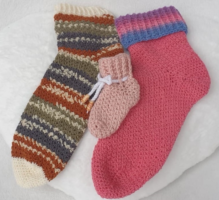 Drei gehäkelte Socken: eine lange gestreifte Socke, eine pinke Socke mit mehrfarbigem Bund und eine kleine rosa Babysocke mit Kordel.