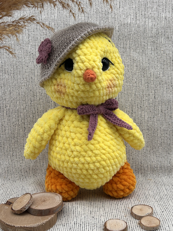 Gehäkeltes gelbes Amigurumi-Küken aus Chenille-Garn mit grauem Hut, lila Schleife, orangefarbenem Schnabel und Füßen vor Stoffhintergrund.