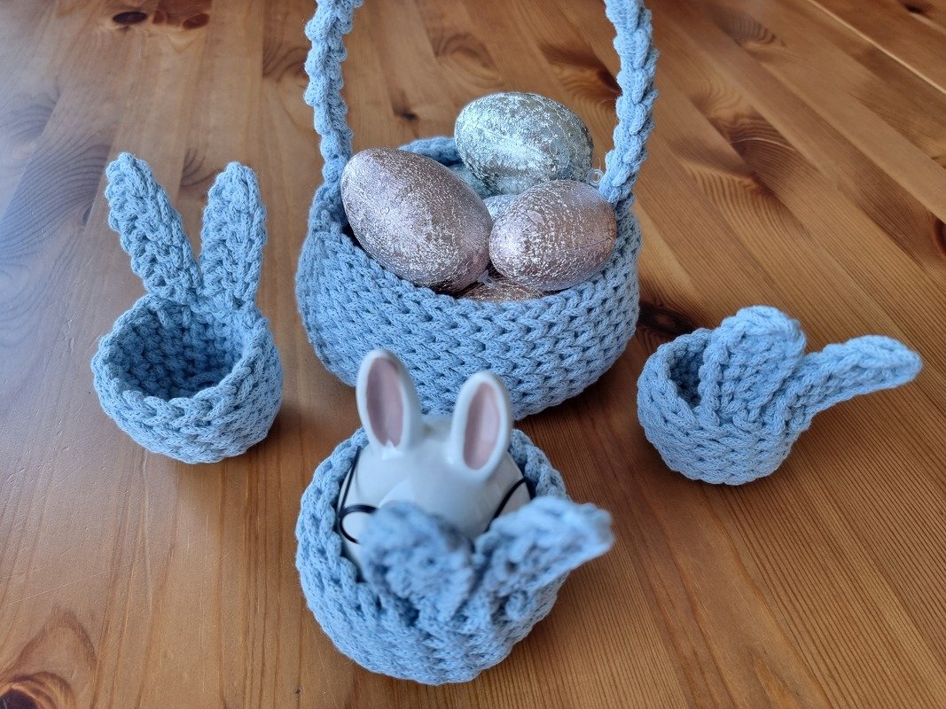 Drei kleine hellblaue gehäkelte Eierkörbchen mit Hasenohren und ein größeres Henkelkörbchen mit Eiern auf Holztisch