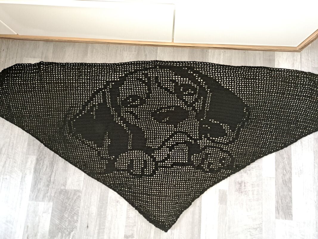 Schwarzes, dreieckiges, gehäkeltes Tuch mit Hundegesicht-Motiv in Filethäkeltechnik auf hellem Boden ausgebreitet.