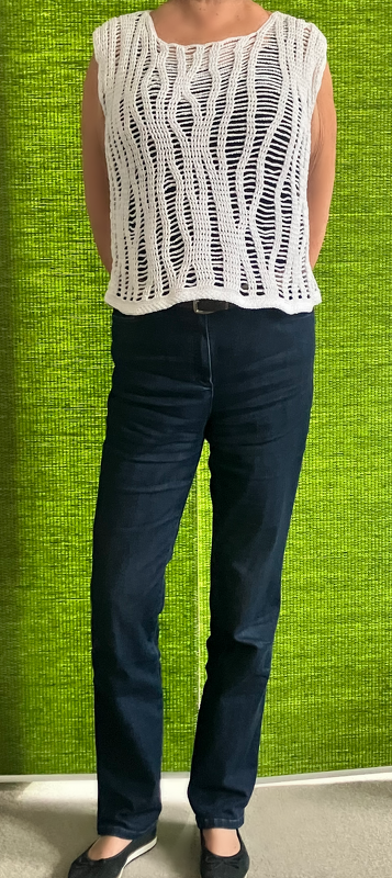 Person trägt weißes, ärmelloses Häkeloberteil mit durchbrochenem Wellen-Ajourmuster über dunklem Top und dunklen Jeans vor grünem Hintergrund.